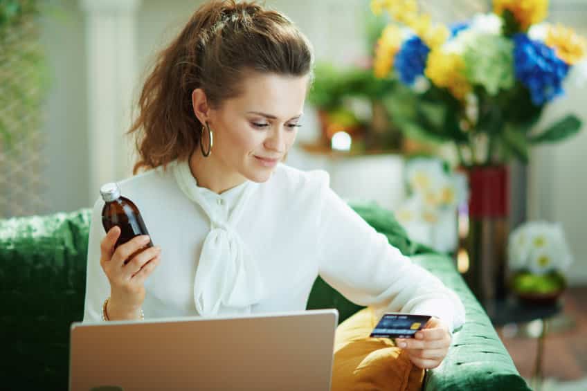 stylish housewife in white blouse with credit card and medicine bottle buying pharmacy on internet using laptop at modern home in sunny day.