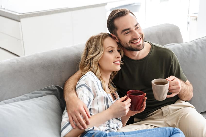 Happy young loving couple indoors at home sitting on sofa hugging drinking coffee or tea watch TV.