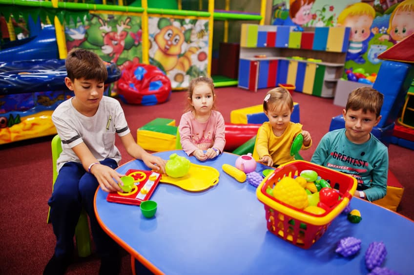 Four kids playing in indoor play center. Kindergarten or preschool play room. Sitting by the table with plastic fruits.