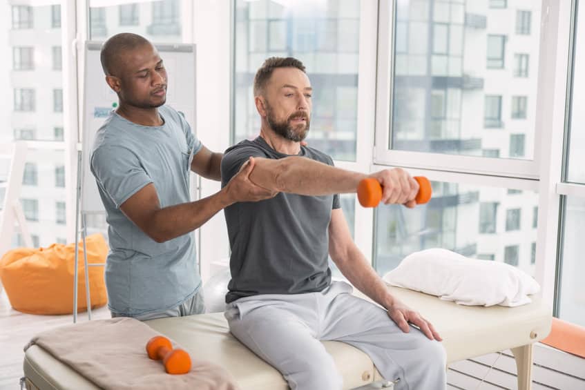 Medical therapist. Pleasant nice doctor standing behind his patient while helping him to raise a hand with a dumbbell